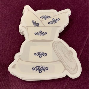 May include: A stack of white and blue floral patterned stickers featuring various kitchenware items, including bowls and a covered dish. The stickers have a white border and are arranged on a dark purple background.