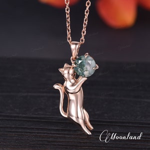 Puede incluir: Un collar de oro rosa con un colgante de gato que sostiene una piedra de ágata verde. El colgante es detallado y el gato está en una pose juguetona.