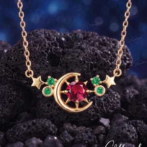 May include: A gold necklace featuring a central crescent moon and star motif, set with a round, deep red gemstone at the center. The design is accented with four small, round green gemstones and two gold stars on either side of the moon.