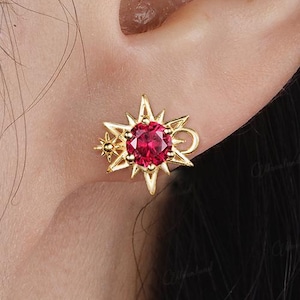 May include: A gold-colored starburst stud earring with a round, red gemstone center. The earring is worn on an ear. The earring has a detailed design with multiple points and smaller star accents. The brand name "Moonland" is in the bottom right corner.