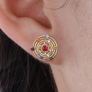 May include: Gold-toned stud earring with a circular design. The earring features a central red gemstone surrounded by concentric circles and colorful gemstones. The earring is worn on an ear.