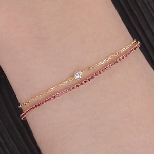 Könnte beinhalten: Zwei zarte goldene Armbänder. Das obere Armband hat einen einzelnen, kleinen, klaren Edelstein. Das untere Armband ist mit einer Reihe kleiner, roter Edelsteine verziert. Die Armbänder sind geschichtet und an einem Handgelenk getragen.