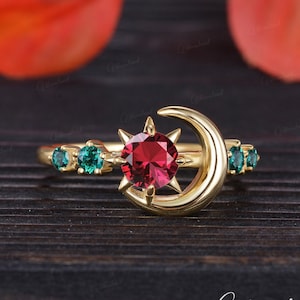 May include: A gold ring with a red gemstone set in a star design and a crescent moon. The ring is accented with smaller green gemstones.