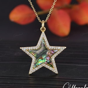 May include: A gold-colored necklace featuring a star-shaped pendant. The pendant has a shimmering, iridescent center and is outlined with small, clear crystals. The necklace is displayed against a dark background with blurred orange and red accents.