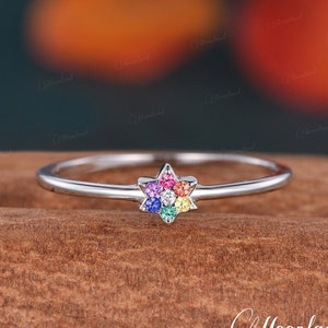 May include: A silver ring featuring a rainbow star-shaped gemstone setting. The ring is displayed on a wooden surface.