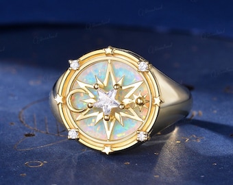 Op de afbeelding: Een gouden ring met een ronde opaal-steen gezet in een kompasontwerp. Het kompas heeft een ster, een halve maan en kleinere sterren rond de rand. De ring is versierd met kleine, heldere stenen.