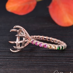May include: A rose gold engagement ring setting with a prong setting and a rainbow colored gemstone band. The band features pink, yellow, and green gemstones.