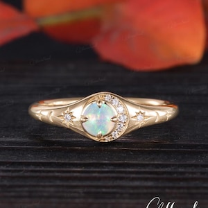 May include: A gold ring featuring a central opal stone surrounded by a halo of small, clear gemstones. The ring's band is adorned with star and crescent moon designs. The ring is set against a dark wooden surface with blurred orange and red foliage in the background.