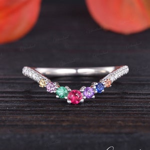 May include: A silver ring with a V-shaped band set with small diamonds and colourful gemstones. The gemstones are arranged in a rainbow pattern, with a red ruby in the centre.