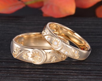 Könnte beinhalten: Zwei goldene Eheringe mit Himmelsmotiven. Ein Ring zeigt ein Mond- und Sternmuster, während der andere Sonnenstrahlen und Diamantakzente aufweist. Die Ringe werden auf einer dunklen Holzoberfläche präsentiert.