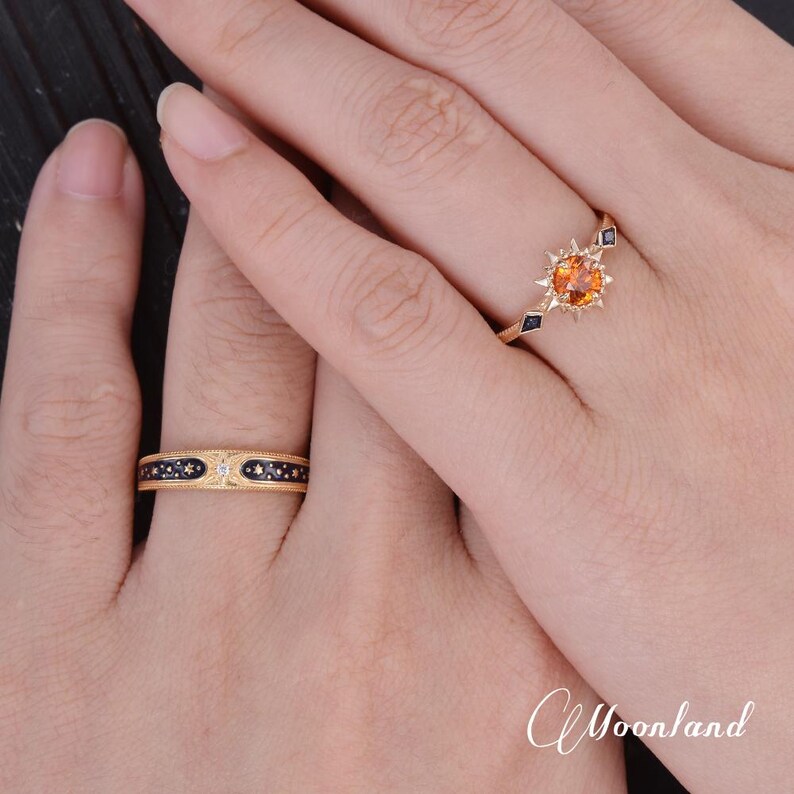 Op de afbeelding: Er worden twee gouden ringen getoond, &eacute;&eacute;n aan elke hand. E&eacute;n ring heeft een centrale oranje edelsteen met een zonnesterontwerp, terwijl de andere een band heeft met een donkerblauw patroon en kleine witte accenten.