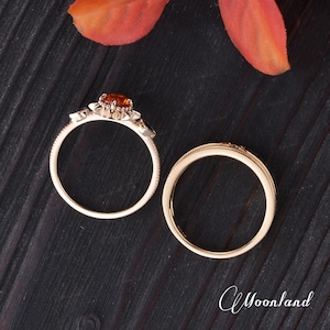 Op de afbeelding: Twee gouden ringen op een donkerhouten oppervlak. E&eacute;n ring heeft een ronde, oranje edelsteen en decoratieve accenten. De andere ring is een eenvoudige band met een textuurontwerp. Een herfstkleurig blad is op de achtergrond.