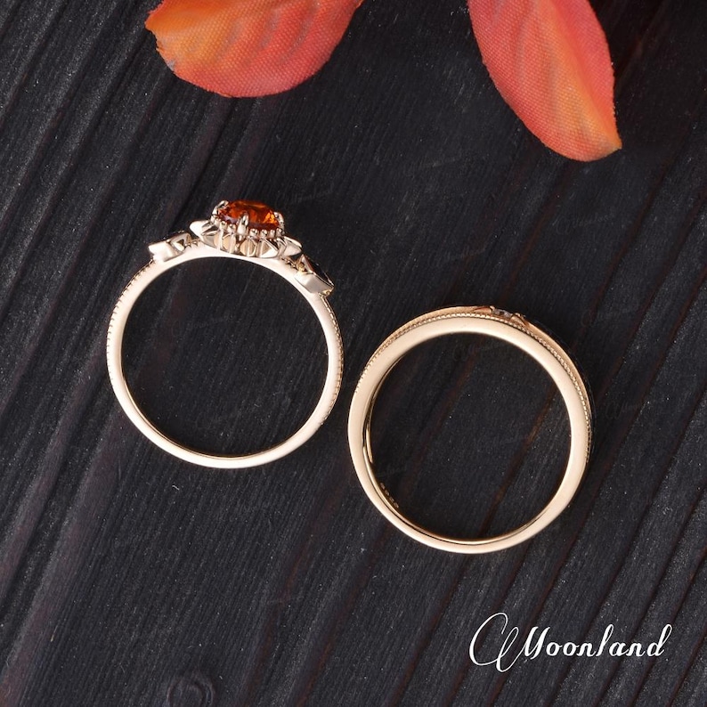 Op de afbeelding: Twee gouden ringen op een donkerhouten oppervlak. E&eacute;n ring heeft een ronde, oranje edelsteen en decoratieve accenten. De andere ring is een eenvoudige band met een textuurontwerp. Een herfstkleurig blad is op de achtergrond.