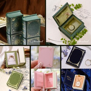 K&ouml;nnte beinhalten: Eine Sammlung kleiner buchf&ouml;rmiger Schmuckk&auml;stchen in verschiedenen Farben wie Gr&uuml;n, Rosa und Dunkelblau. Diese K&auml;stchen verf&uuml;gen &uuml;ber Goldfolienverzierungen, mit Samt ausgekleidete Innenr&auml;ume und enthalten teilweise Schmuckst&uuml;cke.