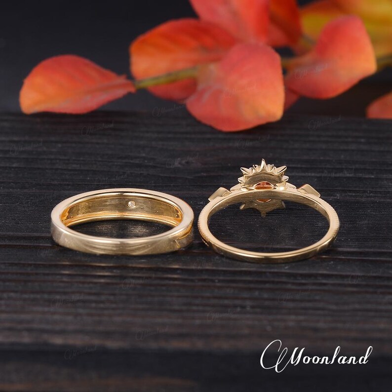 Op de afbeelding: Twee gouden ringen. E&eacute;n is een eenvoudige band met een kleine diamant, de andere heeft een sunburst-ontwerp met een centrale oranje edelsteen. De ringen zijn geplaatst tegen een donkere houten ondergrond met oranje bladeren op de achtergrond.