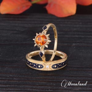 Op de afbeelding: Twee gouden ringen. E&eacute;n ring heeft een grote oranje edelsteen in een sterrenpatroon. De andere ring heeft een donkerblauwe band met gouden sterren en een kleine diamant. De ringen staan tegen een donkere achtergrond.