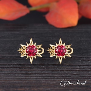 May include: A pair of gold starburst earrings with a red gemstone in the center of each star. The earrings are on a dark wooden surface.