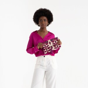 May include: A woman wearing a pink blouse and white pants holds a floral patterned clutch bag with a tassel.