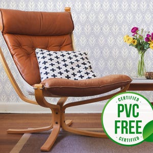 Op de afbeelding: Een bruine leren fauteuil met een zwart-wit gepatroneerd kussen staat voor een wit en blauwe geometrisch gepatroneerde behang. Een vaas met gele en paarse bloemen staat op een houten bijzettafel. Een groen blad met de tekst "Certified PVC Free Certified" is op de voorgrond.