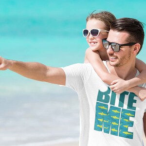 K&ouml;nnte beinhalten: Ein wei&szlig;es T-Shirt mit einem blauen und gelben Aufdruck, auf dem "Bite Me" mit einer Haifischflosse und einem Fischdesign steht. Ein Mann und ein junges M&auml;dchen tragen Sonnenbrillen und l&auml;cheln in die Kamera.