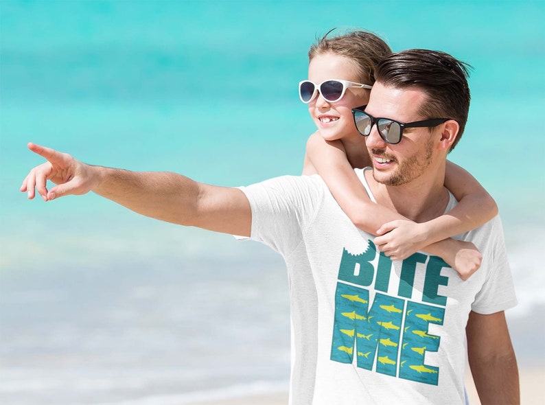 K&ouml;nnte beinhalten: Ein wei&szlig;es T-Shirt mit einem blauen und gelben Aufdruck, auf dem "Bite Me" mit einer Haifischflosse und einem Fischdesign steht. Ein Mann und ein junges M&auml;dchen tragen Sonnenbrillen und l&auml;cheln in die Kamera.