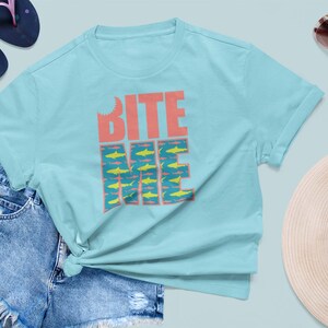 K&ouml;nnte beinhalten: Ein hellblaues T-Shirt mit dem Text "BITE ME" in Pink und Blau. Der Text ist von einem Muster aus gelben Fischen umgeben.