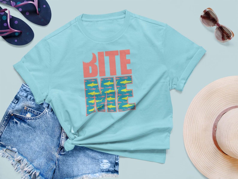K&ouml;nnte beinhalten: Ein hellblaues T-Shirt mit dem Text "BITE ME" in Pink und Blau. Der Text ist von einem Muster aus gelben Fischen umgeben.