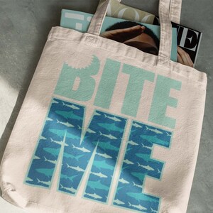K&ouml;nnte beinhalten: Eine wei&szlig;e Canvas-Tragetasche mit dem Text "Bite Me" in blaugr&uuml;nen Buchstaben. Die Buchstaben sind von einem Muster aus blaugr&uuml;nen und hellblauen Haien umgeben.