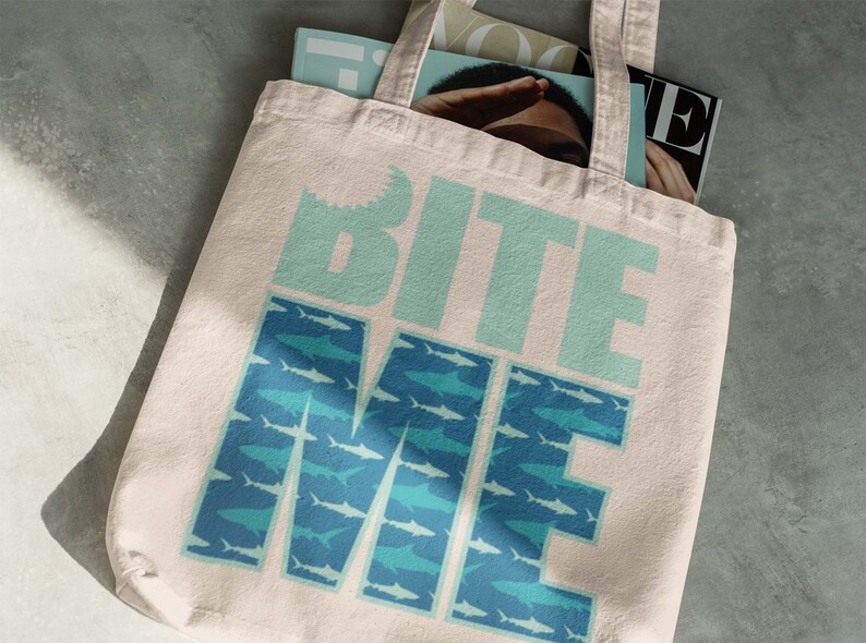 K&ouml;nnte beinhalten: Eine wei&szlig;e Canvas-Tragetasche mit dem Text "Bite Me" in blaugr&uuml;nen Buchstaben. Die Buchstaben sind von einem Muster aus blaugr&uuml;nen und hellblauen Haien umgeben.