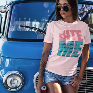 K&ouml;nnte beinhalten: Eine Frau tr&auml;gt ein pinkfarbenes T-Shirt mit dem Text "Bite Me" in Pink und Blau mit einem Hai-Motiv. Sie steht vor einem blauen Vintage-Van.