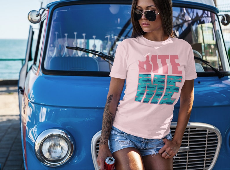 K&ouml;nnte beinhalten: Eine Frau tr&auml;gt ein pinkfarbenes T-Shirt mit dem Text "Bite Me" in Pink und Blau mit einem Hai-Motiv. Sie steht vor einem blauen Vintage-Van.