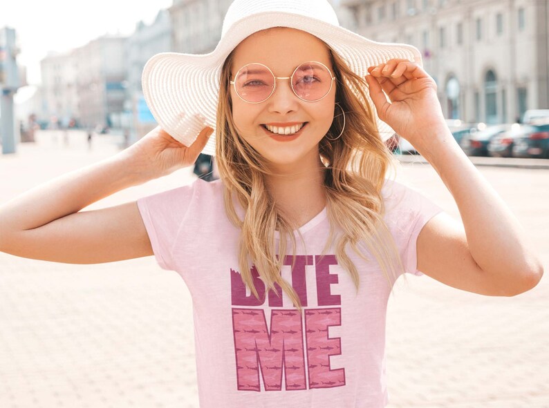 K&ouml;nnte beinhalten: Eine Frau tr&auml;gt ein pinkfarbenes T-Shirt mit dem Text "Bite Me" in pinken Buchstaben. Sie tr&auml;gt einen wei&szlig;en Sonnenhut und runde rosa Sonnenbrillen.