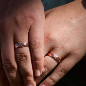 May include: Two hands with matching hammered copper wedding bands. The bands are a warm, reddish-brown color.