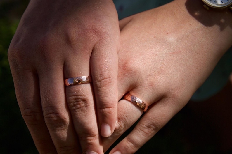 May include: Two hands with matching hammered copper wedding bands. The bands are a warm, reddish-brown color.