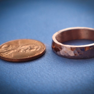 Puede incluir: Un anillo de cobre martillado con una superficie texturizada se encuentra junto a un centavo sobre un fondo azul. El anillo tiene una banda ancha y un dise&ntilde;o facetado. La moneda es una moneda de un centavo estadounidense.