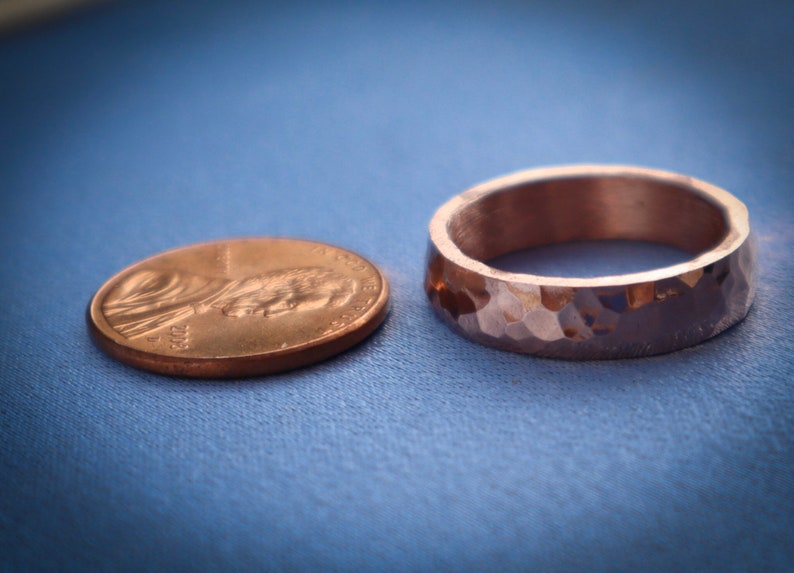 Puede incluir: Un anillo de cobre martillado con una superficie texturizada se encuentra junto a un centavo sobre un fondo azul. El anillo tiene una banda ancha y un dise&ntilde;o facetado. La moneda es una moneda de un centavo estadounidense.