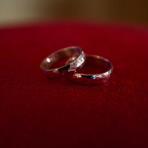 May include: Two silver wedding bands with a hammered texture, resting on a red velvet surface.
