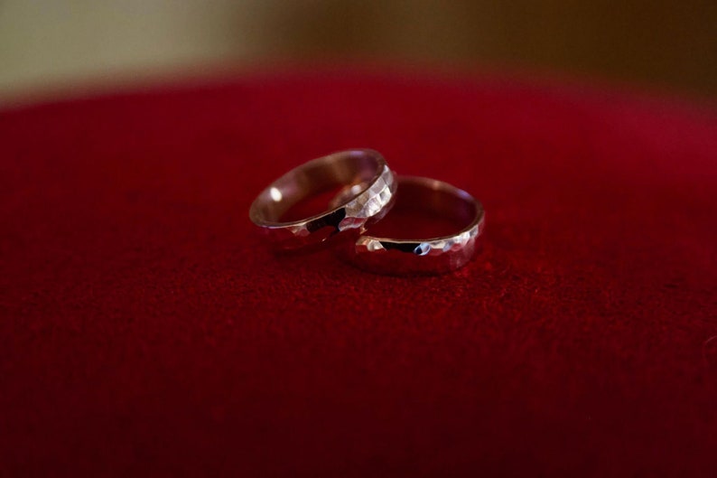 May include: Two silver wedding bands with a hammered texture, resting on a red velvet surface.
