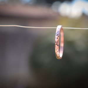 Puede incluir: Un anillo de oro rosa martillado suspendido de un alambre fino. El anillo tiene una superficie texturizada y un dise&ntilde;o simple y elegante. El fondo est&aacute; borroso, con tonos verdes y marrones.