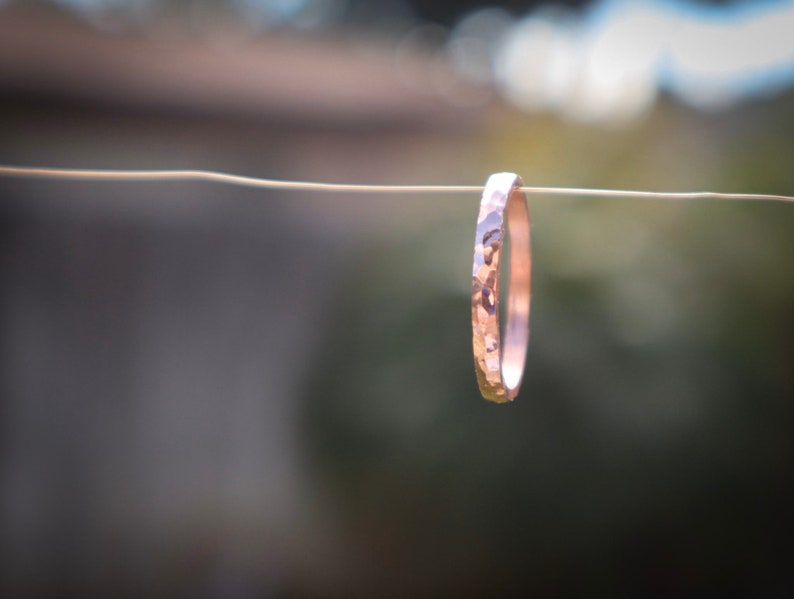 Puede incluir: Un anillo de oro rosa martillado suspendido de un alambre fino. El anillo tiene una superficie texturizada y un dise&ntilde;o simple y elegante. El fondo est&aacute; borroso, con tonos verdes y marrones.