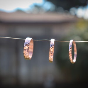 Puede incluir: Tres anillos de cobre martillado cuelgan de un alambre fino sobre un fondo borroso verde y marr&oacute;n. Los anillos tienen una superficie texturizada y un color met&aacute;lico c&aacute;lido. Los anillos son de diferentes anchos, mostrando un dise&ntilde;o de joyer&iacute;a artesanal.