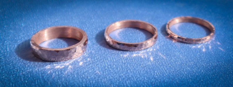 Puede incluir: Tres anillos de metal martillado. Los anillos son de un color met&aacute;lico c&aacute;lido, posiblemente dorado o bronce. Cada anillo tiene una superficie texturizada y facetada, que captura la luz. Los anillos est&aacute;n sobre un fondo azul.