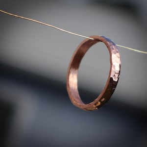 May include: A hammered copper ring with a textured surface. The ring is suspended in the air by a thin wire. The ring's color is a warm, reddish-brown hue, and the surface reflects light.