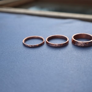 Puede incluir: Tres anillos de cobre martillado de diferentes anchos se muestran sobre una superficie azul. Los anillos tienen un aspecto texturizado y r&uacute;stico, con un tono marr&oacute;n rojizo c&aacute;lido. Los anillos son probablemente hechos a mano.