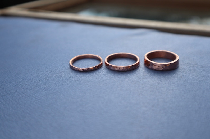 Puede incluir: Tres anillos de cobre martillado de diferentes anchos se muestran sobre una superficie azul. Los anillos tienen un aspecto texturizado y r&uacute;stico, con un tono marr&oacute;n rojizo c&aacute;lido. Los anillos son probablemente hechos a mano.