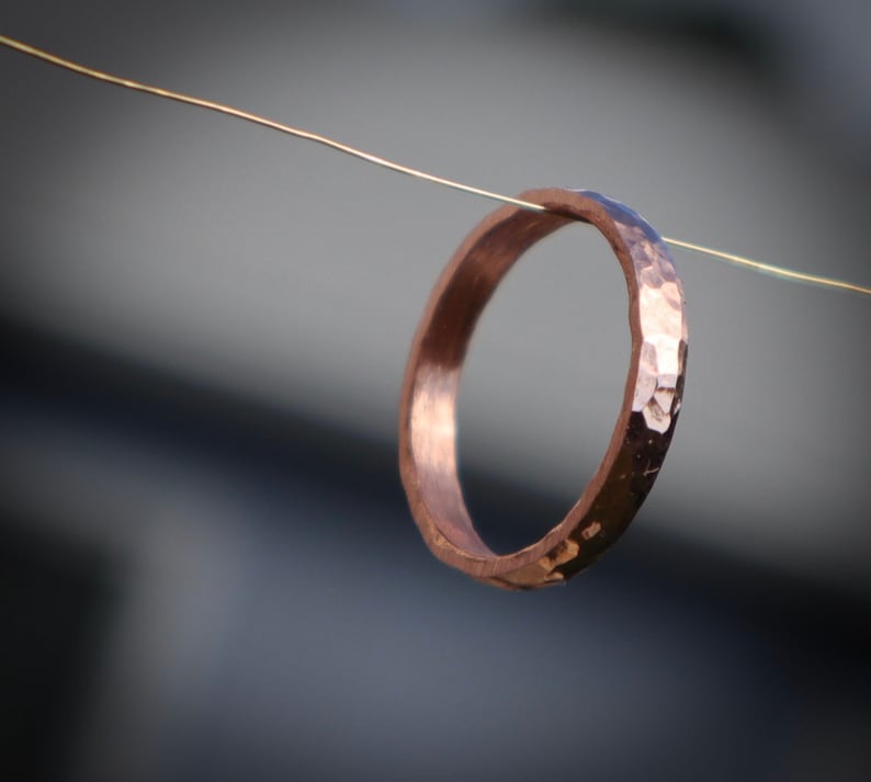 Puede incluir: Un anillo de cobre martillado con una superficie texturizada. El anillo est&aacute; suspendido por un alambre fino. El color del anillo es un tono c&aacute;lido rojizo-marr&oacute;n, y la superficie refleja la luz.