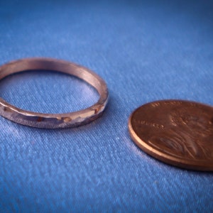 Puede incluir: Un anillo plateado con textura martillada junto a una moneda de cobre sobre tela azul. El anillo es circular y tiene una superficie texturizada. La moneda es ovalada. El di&aacute;metro del anillo es de aproximadamente 1,9 cm.