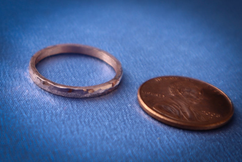 Puede incluir: Un anillo plateado con textura martillada junto a una moneda de cobre sobre tela azul. El anillo es circular y tiene una superficie texturizada. La moneda es ovalada. El di&aacute;metro del anillo es de aproximadamente 1,9 cm.