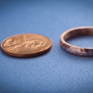 Puede incluir: Un anillo de metal martillado junto a un centavo sobre una superficie azul. El anillo tiene una superficie texturizada y un ancho de banda de aproximadamente 0,25 cm. El centavo es de color cobre y muestra el perfil de una persona.