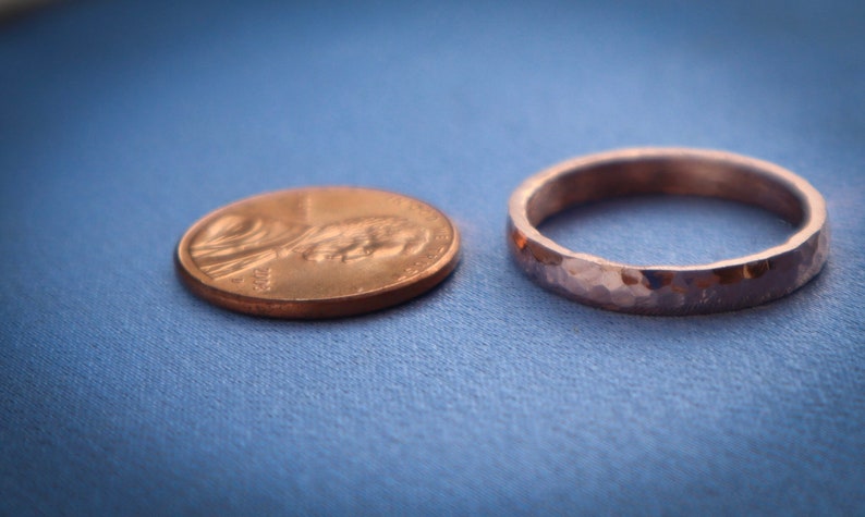 Puede incluir: Un anillo de metal martillado junto a un centavo sobre una superficie azul. El anillo tiene una superficie texturizada y un ancho de banda de aproximadamente 0,25 cm. El centavo es de color cobre y muestra el perfil de una persona.